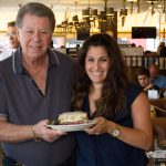 Norm and Interviewer each holding a side of a plate with a sandwich