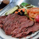 plate full of hot, sliced pastrami with a pickle, olives, tomatoes and other garnishes